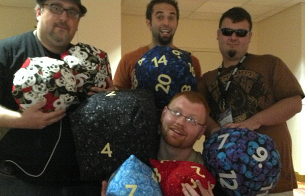 Four unknown GenCon attendees holding the dice pillows as if they had just found the world's greatest treasure. There is a full set of polyhedral dice, including percentage dice, sewn in various quilting cottons that have stars, swirls, and skulls.